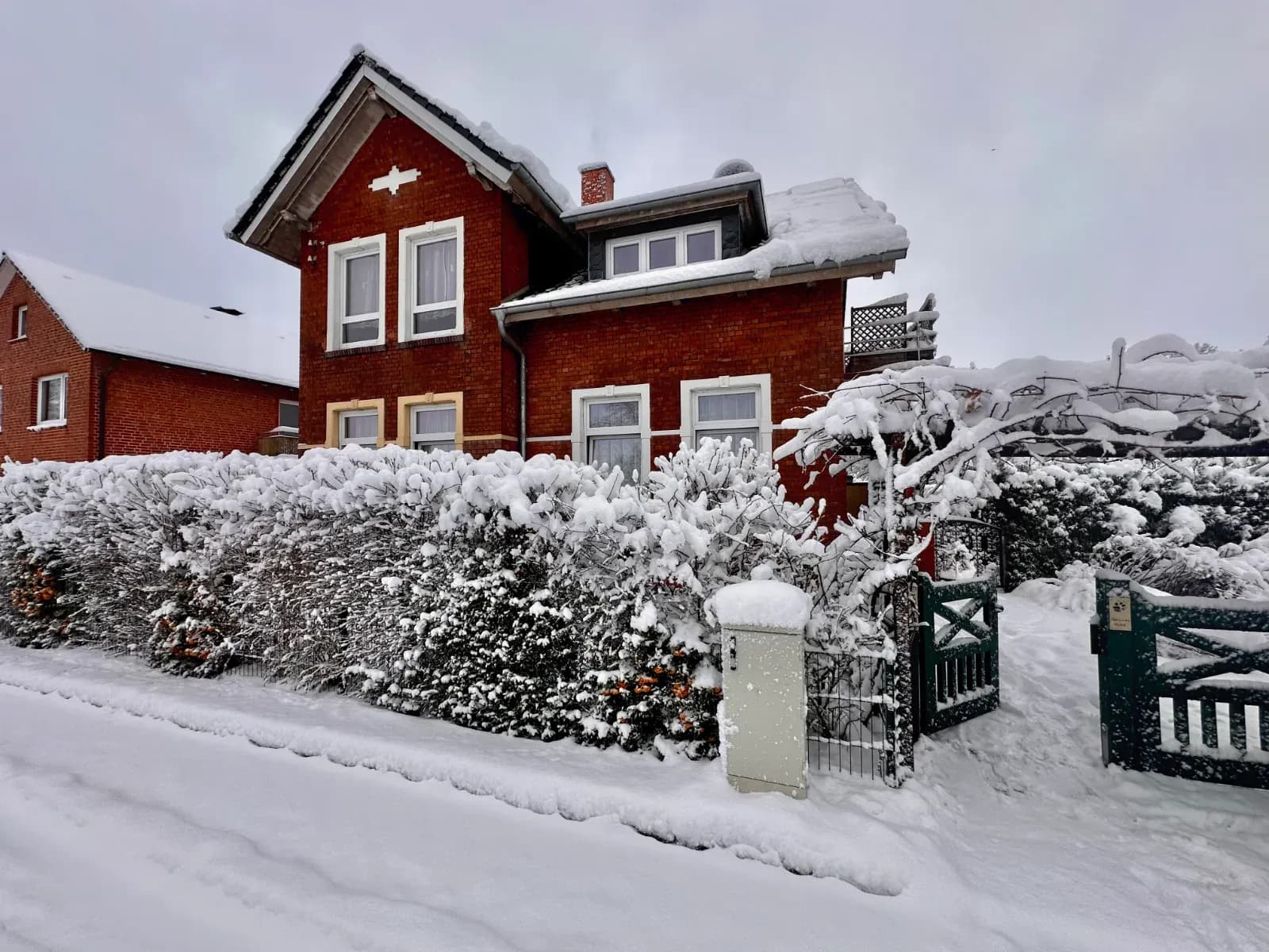Rotes Backsteinhaus hinter einer verschneiten Hecke, davor ein offenes grünes Gartentor