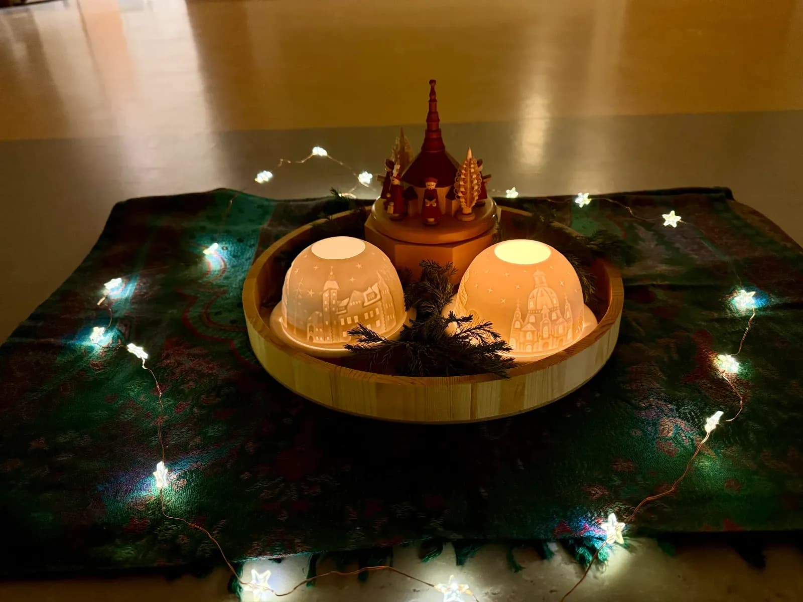 Holztablett mit zwei leuchtenden Porzellan-Windlichtern, einer kleinen Weihnachtspyramide und Tannengrün, darum eine Lichterkette mit Sternen