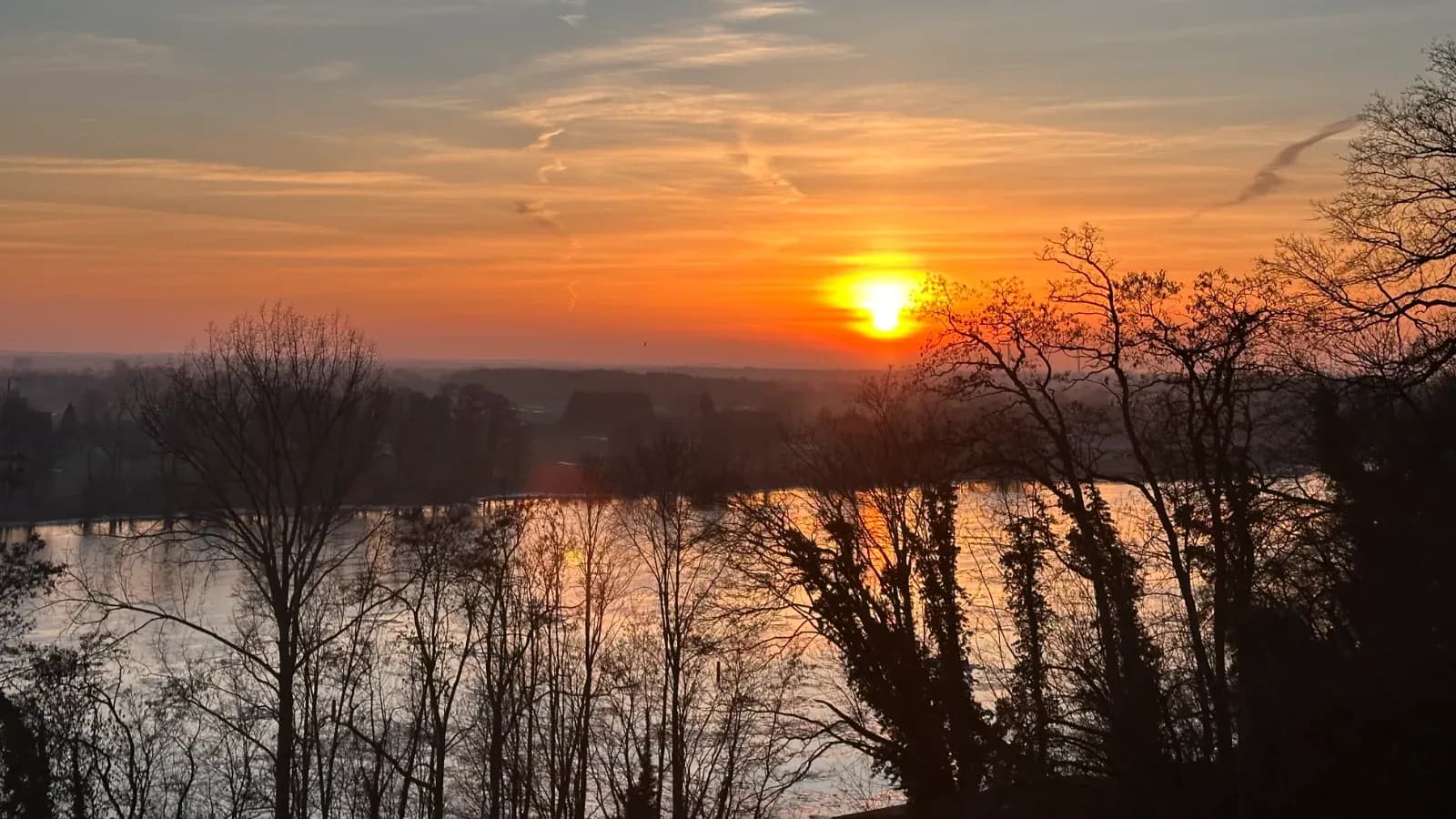 Sonnenuntergang in Orange über einem Fluss, davor kahle Bäume als Silhouetten